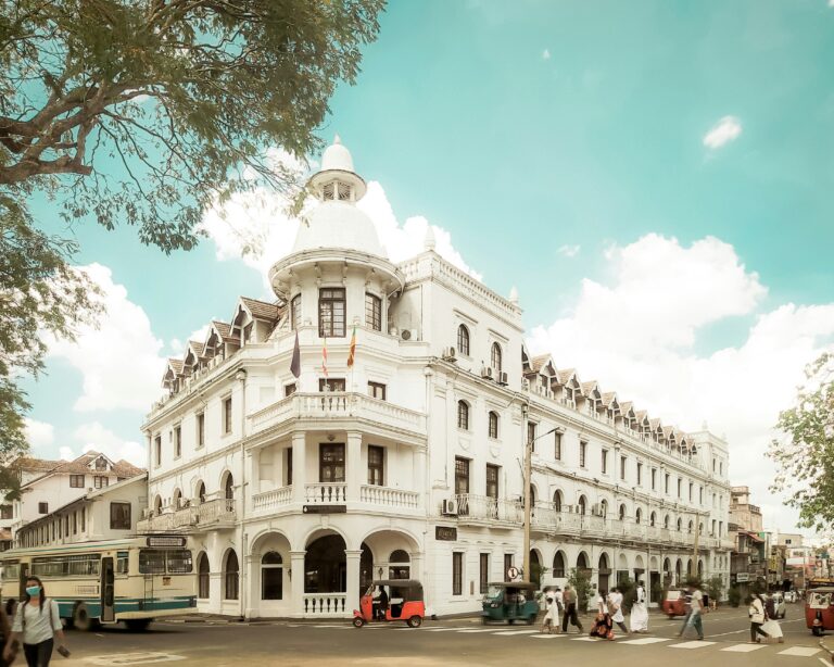 Das weiße, kolonialzeitliche Queen's Hotel an einer Straßenkreuzung im Zentrum von Kandy mit Tuk-Tuks und Bussen während einer Sri Lanka Rundreise.