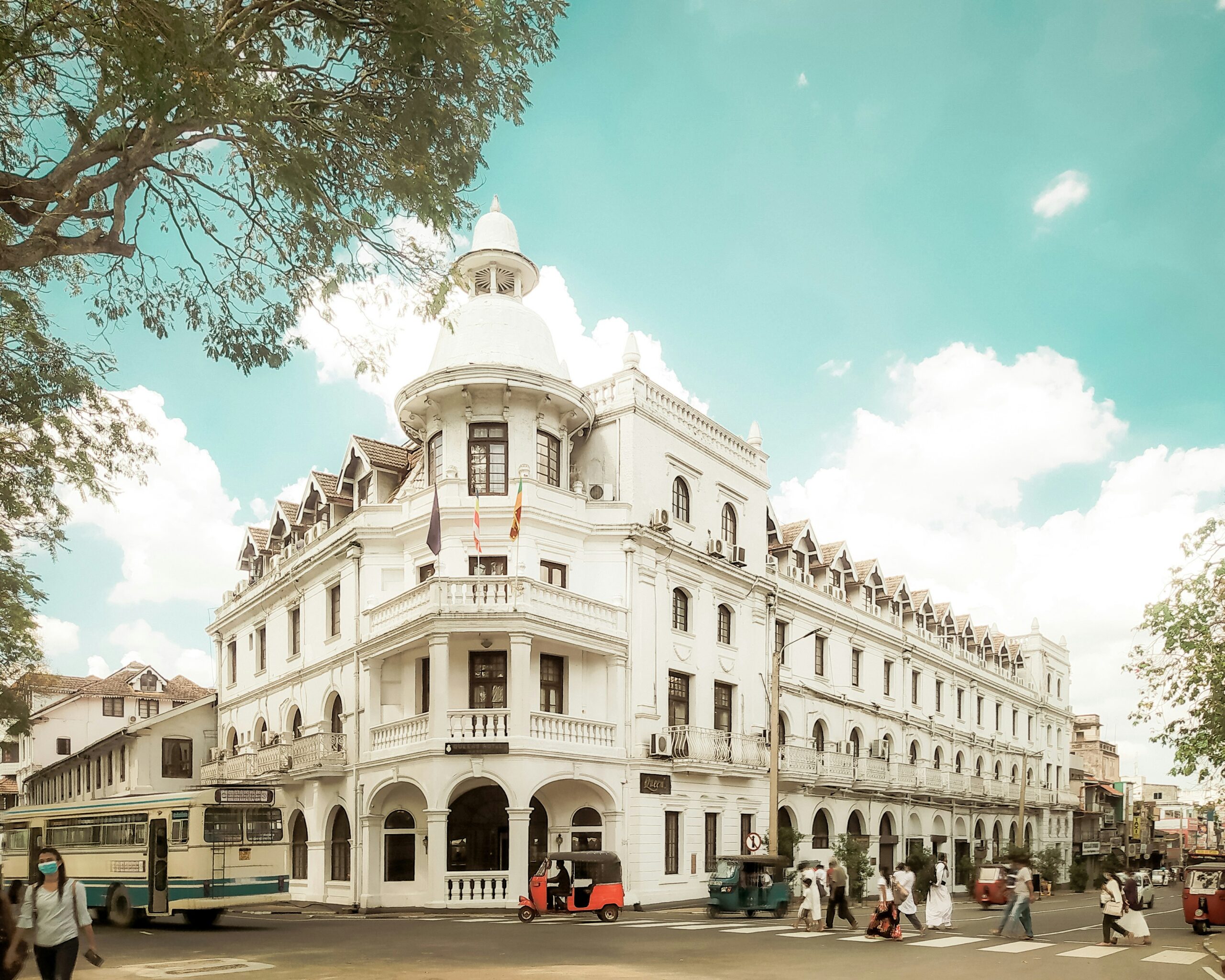 Das weiße, kolonialzeitliche Queen's Hotel an einer Straßenkreuzung im Zentrum von Kandy mit Tuk-Tuks und Bussen während einer Sri Lanka Rundreise.